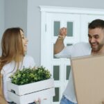 Person sits on the floor of an empty room, surrounded by packed cardboard moving boxes, looking thoughtful about the movin...