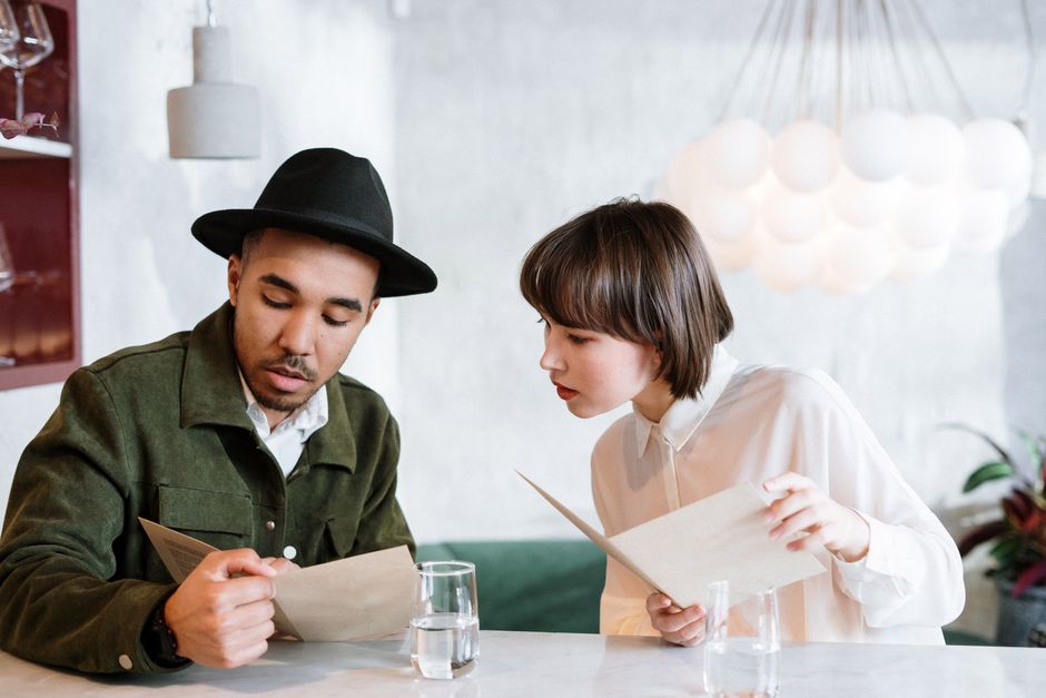 A customer sits at a table in a bright, modern cafe, holding a menu with a pensive and thoughtful expression, deciding what to order.