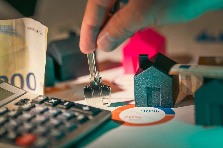 A small model of a house sits on top of a modern calculator against a clean background, representing the financial analysis and cost calculations involved in a home sale.